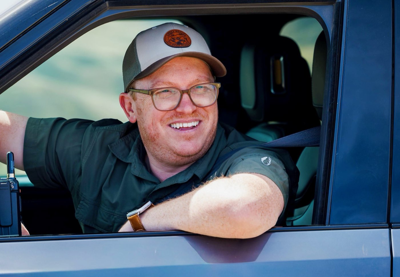 CJ Arnesen smiling and leaning out the open driver's window of his dark blue Rivian truck, wearing glasses and a tan Sierra Nevada Off Road Academy trucker cap, arm resting on the door.