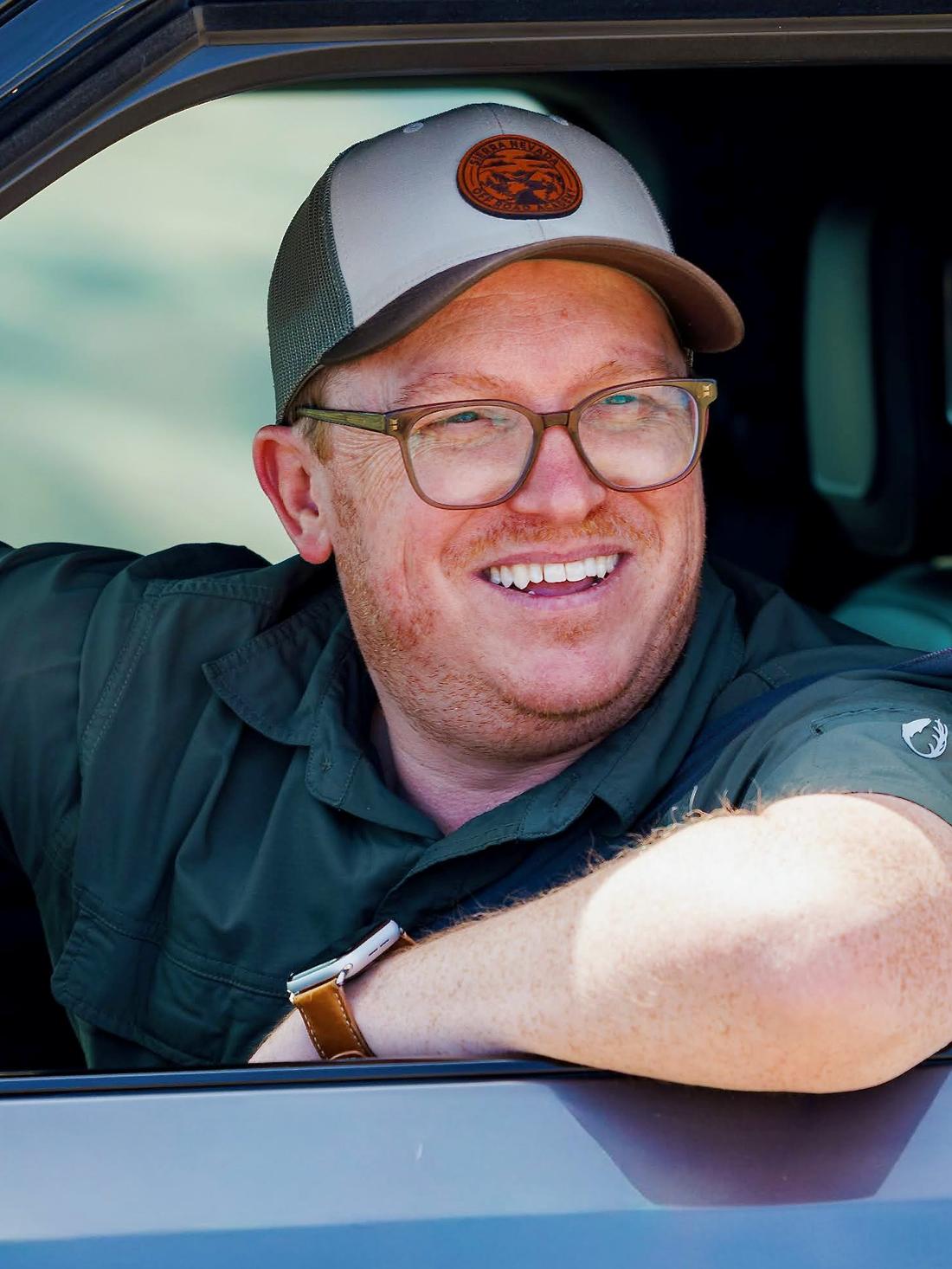 CJ Arnesen smiling from the driver's seat of his Rivian, arm on the window, wearing glasses and a tan Sierra Nevada Off Road trucker cap
