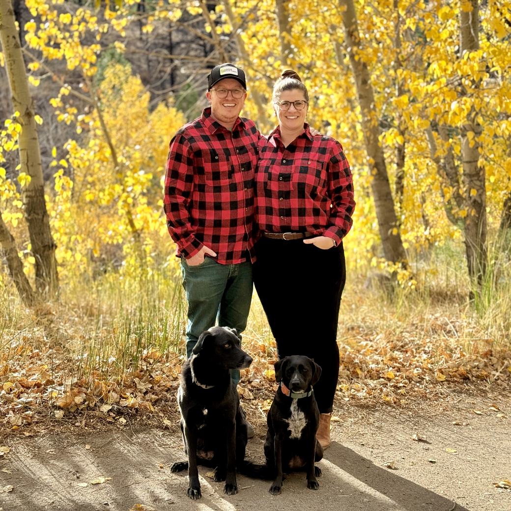 CJ Arnesen and his wife Stefanie standing on a path in the fall aspens, both in red plaid shirts, with their two black dogs sitting in front of them.